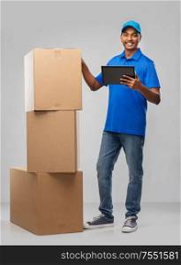 mail service, technology and shipment concept - happy indian delivery man with boxes and tablet computer in blue uniform over grey background. indian delivery man with tablet pc and boxes