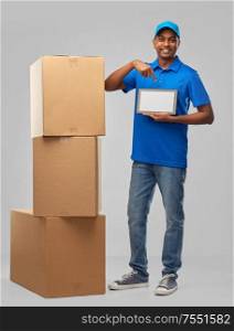 mail service, technology and shipment concept - happy indian delivery man with boxes and tablet computer in blue uniform over grey background. indian delivery man with tablet pc and boxes