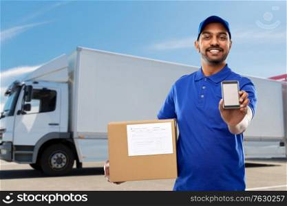 mail service, technology and shipment concept - happy indian delivery man with smartphone and parcel box in blue uniform over truck on street background. indian delivery man with smartphone and parcel box