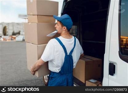 Mailman in uniform unloads the car with parcels, delivery service. Man standing at cardboard packages in vehicle, male deliver, courier or shipping job. Mailman unloads the car with parcels, delivery