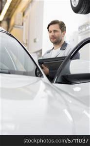 Maintenance engineer holding tablet PC while examining car in repair shop