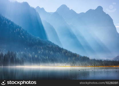 Majestic mountains in bright sunbeams and foggy lake at sunny morning in autumn. Fall in Dolomites, Italy. Landscape with fog over water, high rocks and hills, forest and sunlight at sunrise. Nature. Majestic mountains in bright sunbeams and foggy lake at sunrise