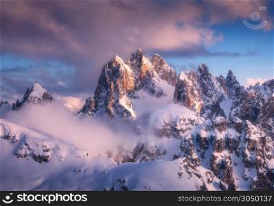 Majestic snowy mountain peaks in fog and blue sky with purple clouds at sunset. Winter landscape with beautiful snow covered rocks in low clouds in frosty evening. Nature in Dolomites, Italy. Alps