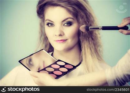 Make up and beauty. Makeup artist applying with brush powder rouge on female check, holding palette with different color pigments, ob blue. Makeup artist applying with brush rouge on female check