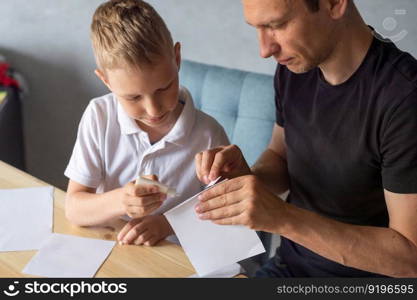 make with your hands, assemble origami, glue parts, cut paper, detail, dad and son, glue, cardboard, white detail, cut out of paper, glue drop, scissors, with your own hands, handmade, daddy&rsquo;s help. A cute boy is sitting with his dad at the table and collecting origami. Holds a white sheet of paper