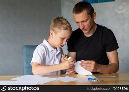 make with your hands, assemble origami, glue parts, cut paper, detail, dad and son, glue, cardboard, white detail, cut out of paper, glue drop, scissors, with your own hands, handmade, daddy’s help. A cute boy is sitting with his dad at the table and collecting origami. Holds a white sheet of paper