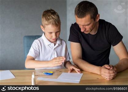 make with your hands, assemble origami, glue parts, cut paper, detail, dad and son, glue, cardboard, white detail, cut out of paper, glue drop, scissors, with your own hands, handmade, daddy&rsquo;s help. A cute boy is sitting with his dad at the table and collecting origami. Adds up the details