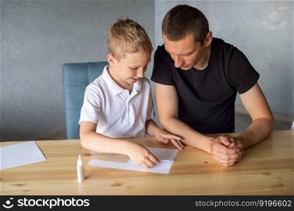 make with your hands, assemble origami, glue parts, cut paper, detail, dad and son, glue, cardboard, white detail, cut out of paper, glue drop, scissors, with your own hands, handmade, daddy&rsquo;s help. A cute boy is sitting with his dad at the table and collecting origami. Glue the parts