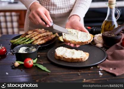 Making breakfast toasts - woman smearing cream cheese on a grilled bread over kitchen table with Asparagus wrapped with bacon and spices on a plate.. Making breakfast toasts - woman smearing cream cheese on a grilled bread over kitchen table with Asparagus wrapped with bacon and spices on a plate