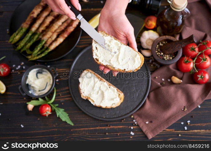 Making breakfast toasts - woman smearing cream cheese on a grilled bread over kitchen table with Asparagus wrapped with bacon and spices on a plate.. Making breakfast toasts - woman smearing cream cheese on a grilled bread over kitchen table with Asparagus wrapped with bacon and spices on a plate