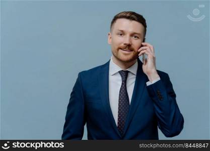 Making business call. Smiling businessman in suit talking on mobile phone with client or partner while standing isolated over light steel blue background, male entrepreneur enjoying conversation. Happy businessman enjoying conversation on mobile phone
