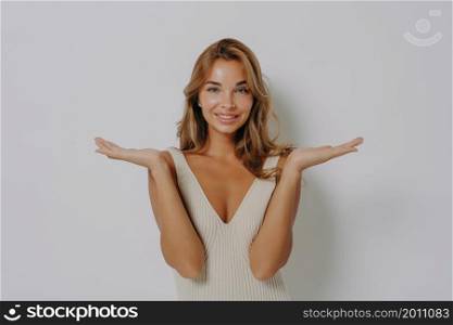 Making decision. Positive slightly confused with nice smile blonde young woman raises hands and spreads palms in hesitation with doubt about question, wears tight dress, isolated on grey background. Positive slightly confused with nice smile blonde young woman raises hands in hesitation