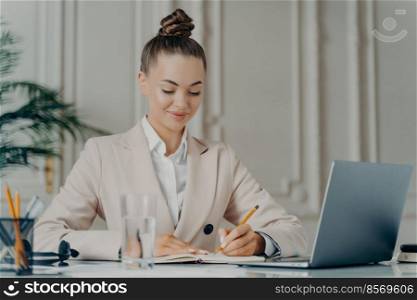 Making to do list. Successful businesswoman in elegant suit writing down points in notepad while sitting at home office, planing working day. Happy female employee in formal wear enjoying work. Attracative successful businesswoman in elegant suit making to do list at work