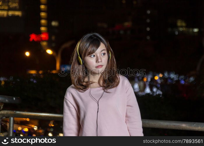 Malaysian female wearing yellow headphones and holding cell phone with city lights in background