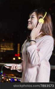 Malaysian female wearing yellow headphones and holding cell phone with city lights in background