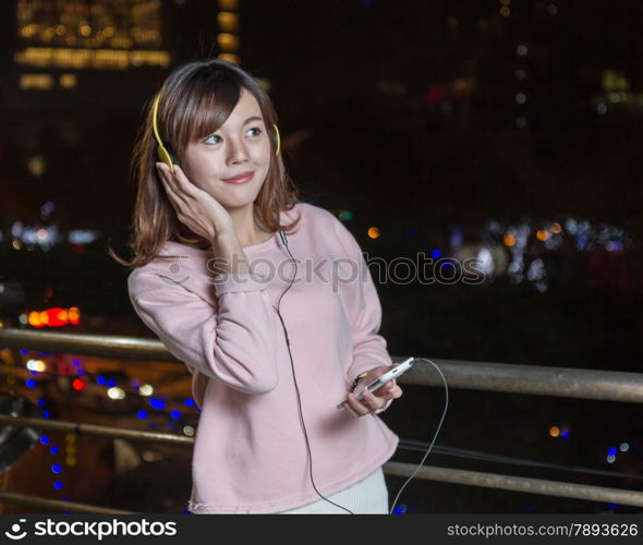 Malaysian female wearing yellow headphones and holding cell phone with city lights in background