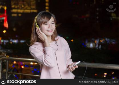 Malaysian female with braces wearing yellow headphones and holding cell phone with city lights in background