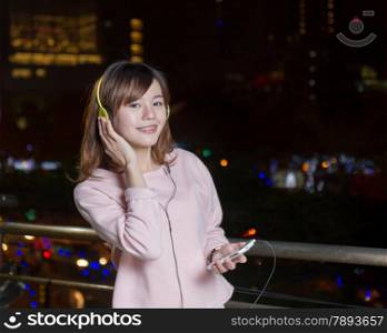 Malaysian female with braces wearing yellow headphones and holding cell phone with city lights in background