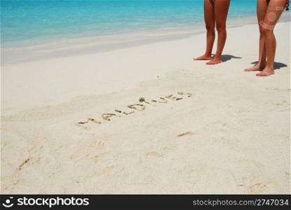 Maldives note written on a white sandy beach with tanned women legs