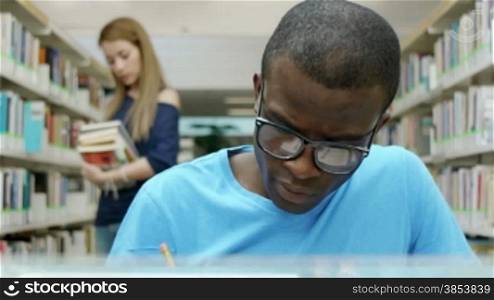 Male african american college student reading book in library, with caucasian woman taking book from shelves in background. Dolly shot