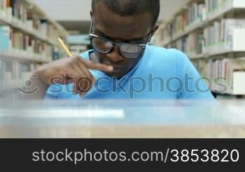 Male african american college student sitting in library, reading book and taking notes.