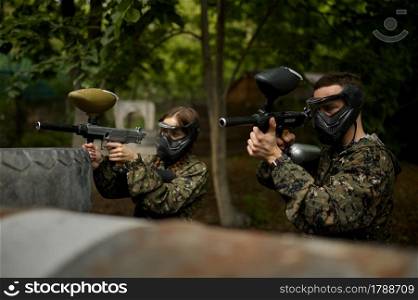 Male and famale warriors in camouflages and masks aimimg with paintball guns. Extreme sport with pneumatic weapon and paint bullets or markers, military team game outdoors, combat tactics. Warriors aimimg with paintball guns, team game
