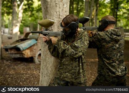 Male and famale warriors in camouflages and masks shooting with paintball guns. Extreme sport with pneumatic weapon and paint bullets or markers, military team game outdoors. Warriors shooting with paintball guns, team game