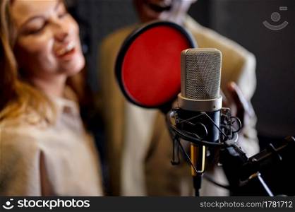 Male and female singers in headphones sings a song at micriphone, recording studio interior on background. Professional voice record, musician workplace, creative process, modern audio technology. Male and female singers sings, recording studio