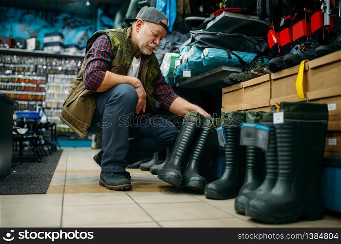 Male angler choosing rubber boots in fishing shop. Equipment and tools for fish catching and hunting. Male angler choosing rubber boots in fishing shop