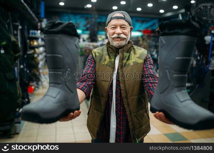 Male angler choosing rubber boots in fishing shop. Equipment and tools for fish catching and hunting. Male angler choosing rubber boots in fishing shop