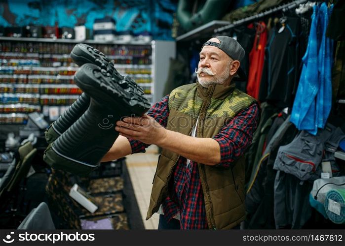 Male angler choosing rubber boots in fishing shop. Equipment and tools for fish catching and hunting, accessory choice on showcase in store. Male angler choosing rubber boots in fishing shop