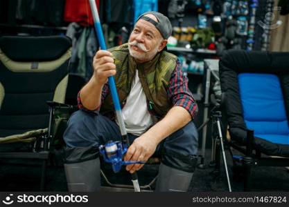 Male angler in rubber boots holds rod in fishing shop, first-person view. Equipment and tools for fish catching and hunting, accessory choice on showcase in store. Angler in rubber boots holds rod in fishing shop