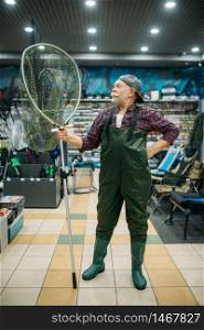 Male angler in rubber jumpsuit holds net in fishing shop, hooks and baubles on background. Fisherman buying equipment and tools for fish catching and hunting, assortment on showcase in store. Angler in rubber jumpsuit holds net, fishing shop
