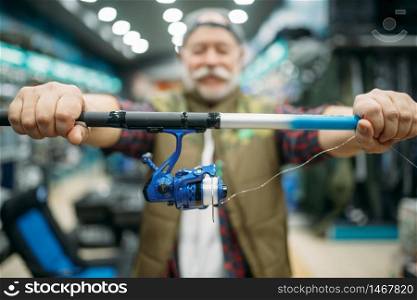 Male angler shows rod with reel in fishing shop. Equipment and tools for fish catching and hunting, accessory choice on showcase in store, spinnings and telescopes assortment. Male angler shows rod with reel in fishing shop