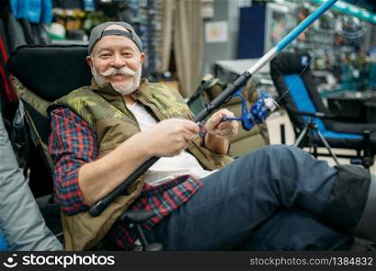 Male angler with rod sitting in chair, fishing shop. Equipment and tools for fish catching and hunting, accessory choice on showcase in store, spinnings and telescopes assortment. Angler with rod sitting in chair, fishing shop
