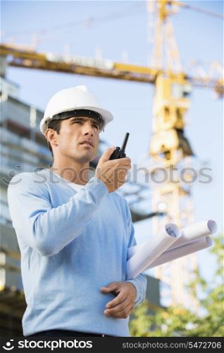 Male architect holding blueprints while using walkie-talkie at construction site