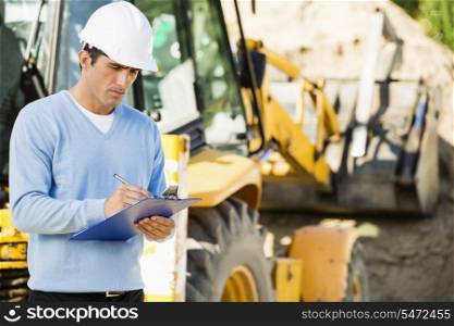 Male architect writing on clipboard against earthmover at construction site