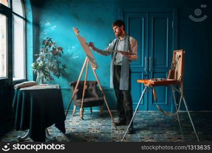 Male artist with palette and brush in hand paint on easel in front of window. Oil paint, paintbrush drawing