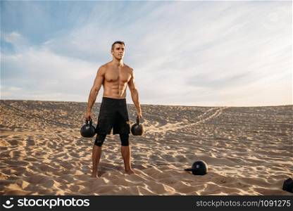Male athlete doing exercises with two kettlebell in desert at sunny day. Strong motivation in sport, strength outdoor training. Male athlete with two kettlebells in desert