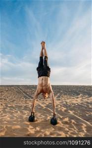 Male athlete doing exercises with two kettlebells stands on hands in desert at sunny day. Strong motivation in sport, strength outdoor training