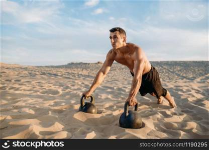 Male athlete doing push-up exercises with two kettlebells in desert at sunny day. Strong motivation in sport, strength outdoor training. Male athlete doing push-up exercises in desert