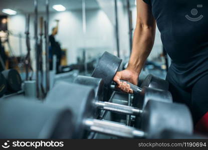 Male athlete takes heavy dumbbell in hand, gym interior on background. Weightlifting workout in sport or fitness club, weight choosing concept