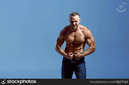 Male athlete with muscular body shows his biceps in studio, blue background. One man with athletic build, shirtless sportsman in jeans pants, active healthy lifestyle. Athlete with muscular body shows biceps in studio