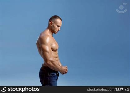 Male athlete with muscular body shows his biceps in studio, side view, blue background. One man with athletic build, shirtless sportsman in jeans pants, active healthy lifestyle. Male athlete shows his biceps in studio, side view