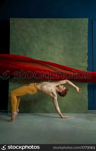 Male ballet dancer doing exercise in dancing studio, grunge wall and red cloth on background. Performer with muscular body, grace and elegance of movements. Male ballet dancer doing exercise, dancing studio