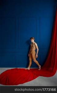 Male ballet dancer poses at blue wall in dancing studio, red cloth on background. Performer with muscular body, grace and elegance of movements. Ballet dancer poses at blue wall in dancing studio
