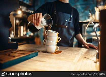 Male barista pours black coffee on a stack of cups, cafe counter and espresso machine on background. Barman works in cafeteria, bartender occupation. Male barista pours black coffee on a stack of cups