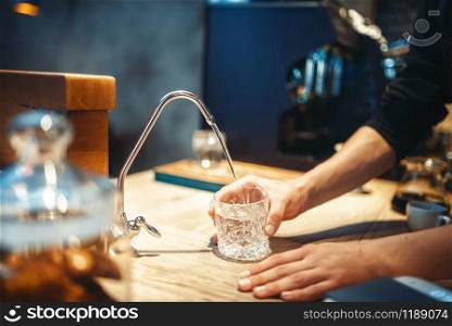 Male barista pours filtered water into the glass, cafe or bar counter on background. Professional coffee preparation by bartender in cafeteria, barman occupation. Male barista pours filtered water into the glass