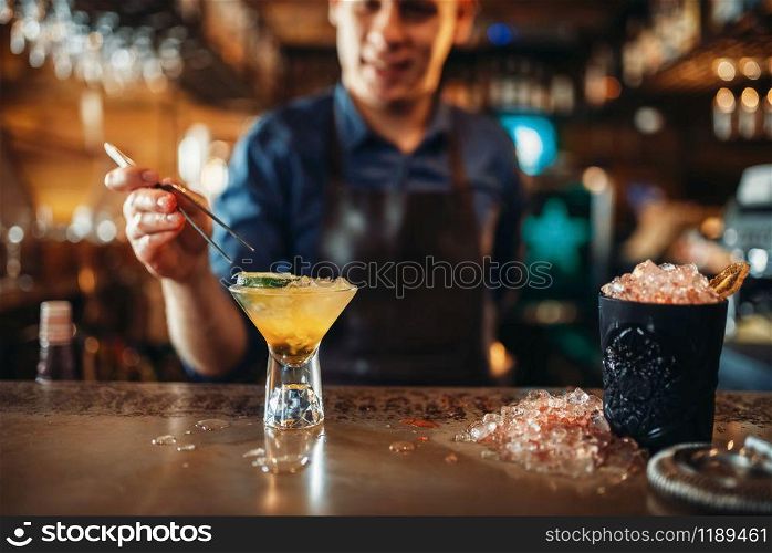 Male barman making coctail with the help of forceps. Barkeeper occupation, bartender at the bar counter. Barman making coctail with the help of forceps