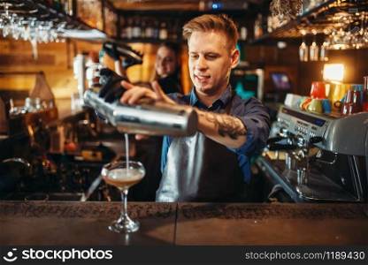 Male barman pouring the drink from the shaker at the bar counter. Alcoholic coctail preparation. Barkeeper occupation, bartending. Male barman pouring the drink from the shaker